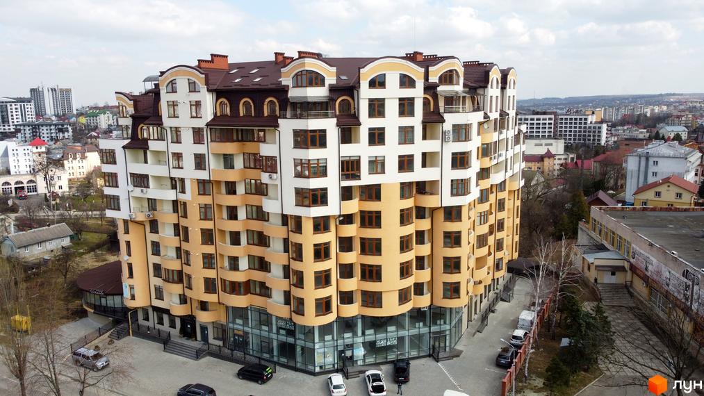 Новобудова на вул. Роксолани, 16, з багатоярусним фасадом білого та жовтого кольорів, з вигнутими елементами та коричневим дахом. Навколо будівлі розташована прибудинкова територія з парковкою та деревами.