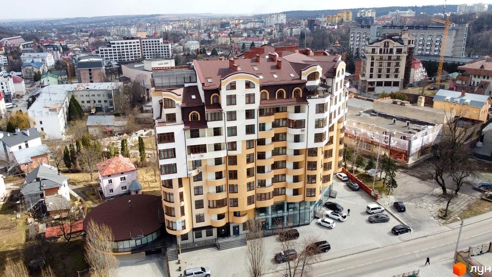 Висотна будівля житлового комплексу з фасадом у світлих тонах. На прибудинковій території розташована парковка для автомобілів.