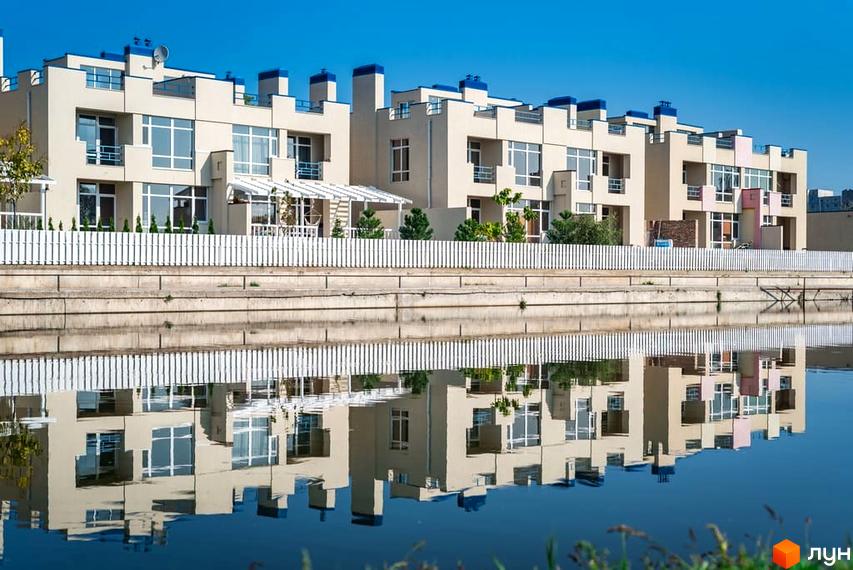 Зображення житлового комплексу Lake City. Будинки з великими вікнами та балконами, біла огорожа та відображення у воді, блакитне небо.