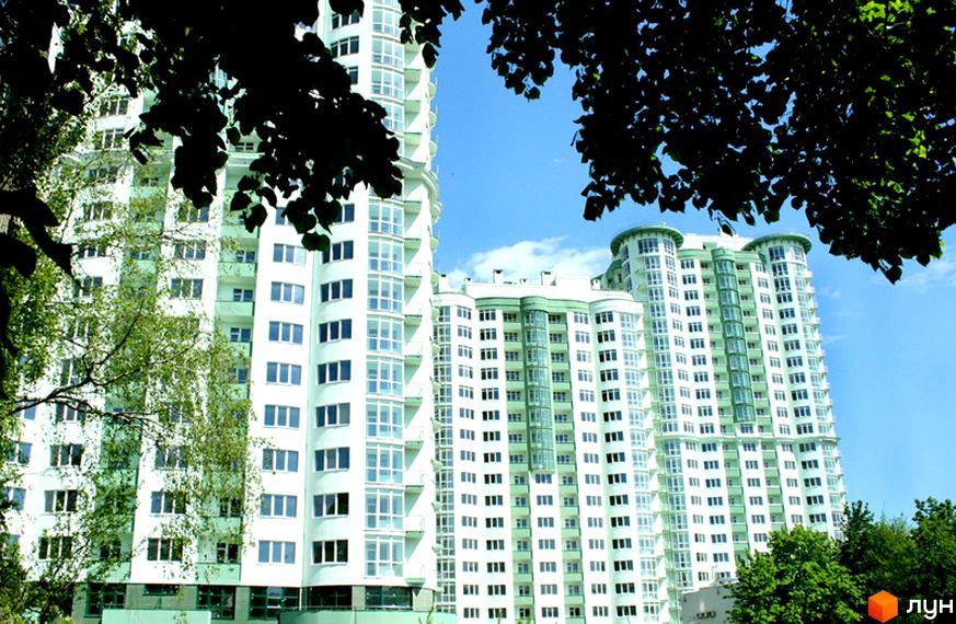 Житловий комплекс Смарагдовий: багатоповерхові будинки білого кольору з зеленими елементами та округлими балконами. Територія оточена деревами та зеленню.