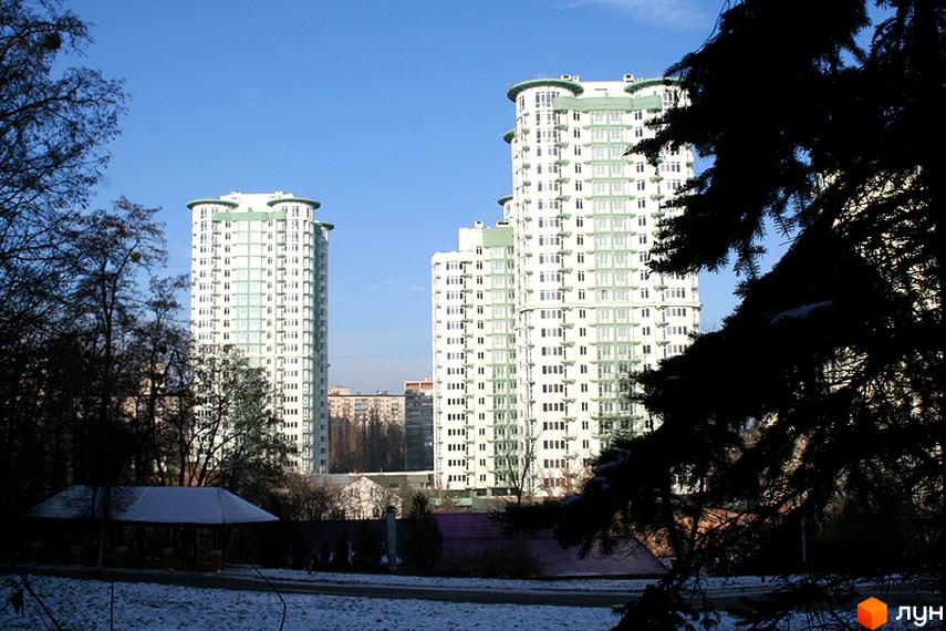 Високі білі житлові будинки Житлового комплексу Смарагдовий зі світло-зеленими акцентами та округлими дахами. На передньому плані засніжений парк з деревами та альтанкою.