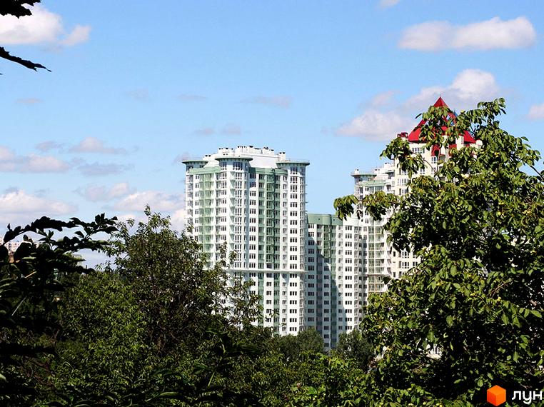 Житловий комплекс Смарагдовий представлений високими будинками світлого кольору з зеленими акцентами. Навколо будівель ростуть густі зелені дерева.