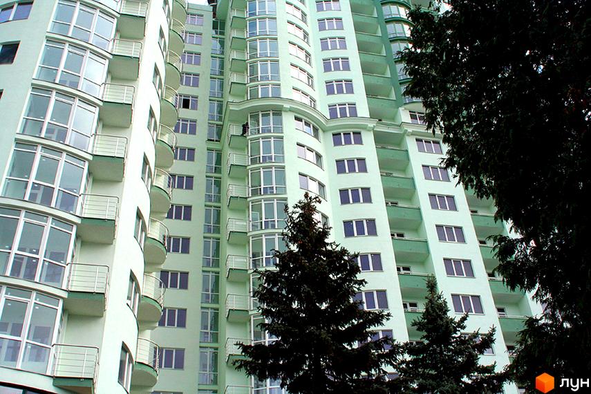 Житловий комплекс Смарагдовий: багатоповерховий будинок світло-зеленого кольору з численними балконами та великими вікнами. Прибудинкова територія озеленена хвойними деревами.