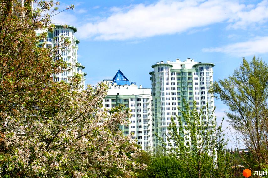 Багатоповерхові будинки Житлового комплексу Смарагдовий мають біло-зелені фасади та округлі секції. Перед ними розташовані квітучі дерева та зелені насадження.