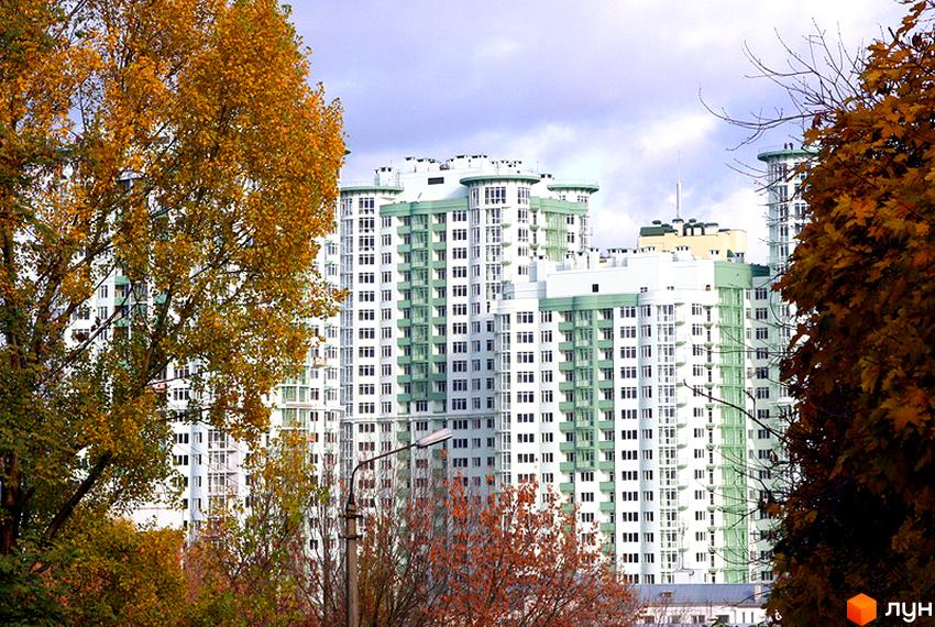 Житловий комплекс Смарагдовий з високими білими будинками та зеленими елементами на фасаді, оточений осінніми деревами.