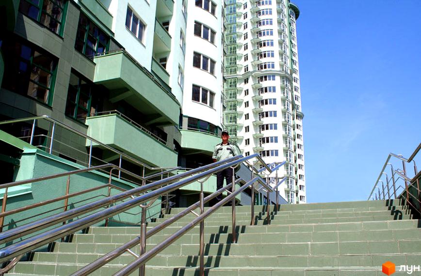 Житловий комплекс Смарагдовий: високий будинок білого кольору з балконами та прилегла будівля із зеленими фасадами. Широкі сходи з металевими поручнями ведуть до входу.