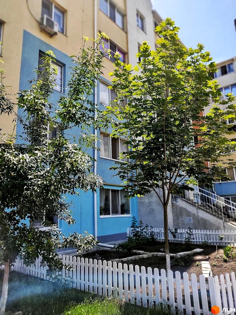 Зовнішній вигляд будинку ЖК Озерки з блакитним фасадом та зеленими деревами на прибудинковій території, огородженій білим парканом.