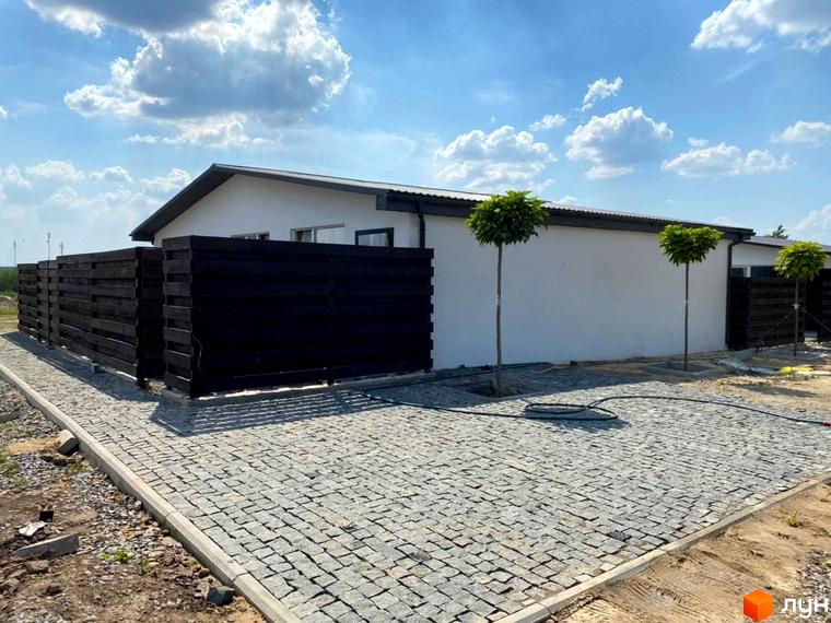 Одноповерховий будинок з білим фасадом, темним дахом та дерев'яним парканом. Прибудинкова територія викладена бруківкою, ростуть три дерева.