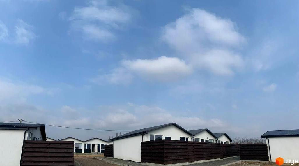 Таунхаус Белгород - це сучасний житловий комплекс з білими фасадами та темними дахами, оточений дерев'яними парканами. Навколо розташовані зелені насадження та доглянута територія.