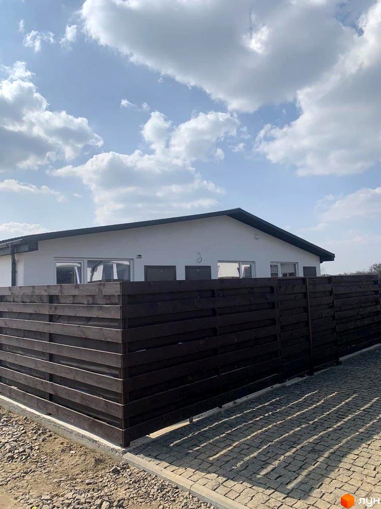 Зовнішній вигляд таунхауса Белгород з темним дерев'яним парканом та бруківкою на прибудинковій території. Бельгійський комплекс з білою стіною та вікнами.