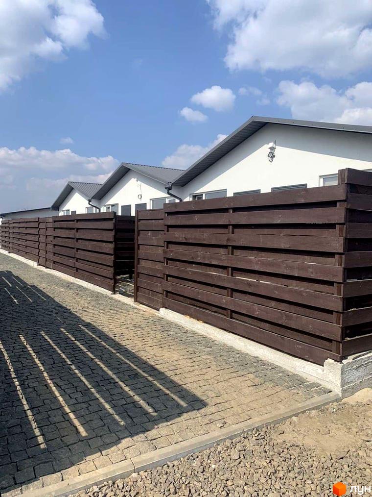 Таунхаус Белгород з коричневим дерев'яним парканом та бруківкою на прибудинковій території. Будинки з сірими дахами на фоні блакитного неба з хмарами.