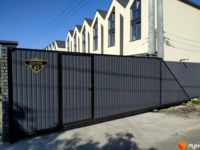 Таунхауси „На Дачній“: сірий металевий паркан з золотистою табличкою №62 та цегляними стовпами. На задньому плані видно частину світлої будівлі таунхаусів під блакитним небом.