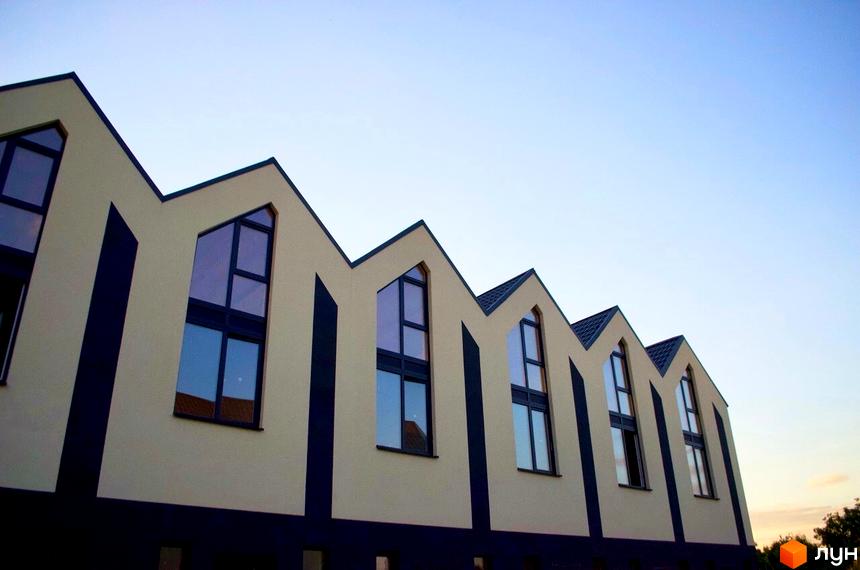 Зовнішній вигляд новобудови Таунхауси „На Дачній“ з характерним зигзагоподібним дахом та великими вікнами на тлі блакитного неба. Будинок має світло-бежевий фасад з темними вертикальними елементами.