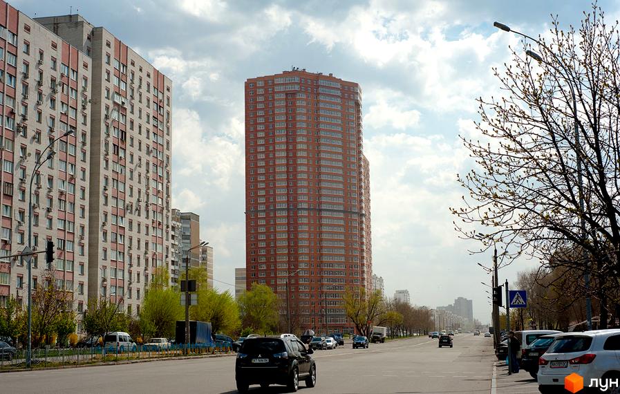 Житловий комплекс Ревуцького, 9: висока цегляна новобудова з балконами, поруч дорога з автомобілями та дерева.