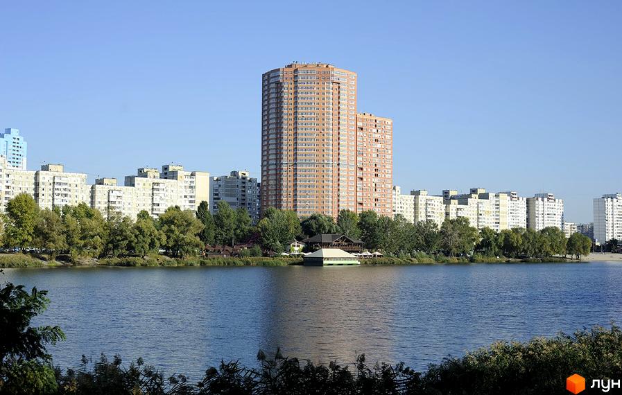 Житловий комплекс Ревуцького, 9 з висоти, оточений зеленими насадженнями та водоймою. На передньому плані видно прибудинкову територію з альтанкою.