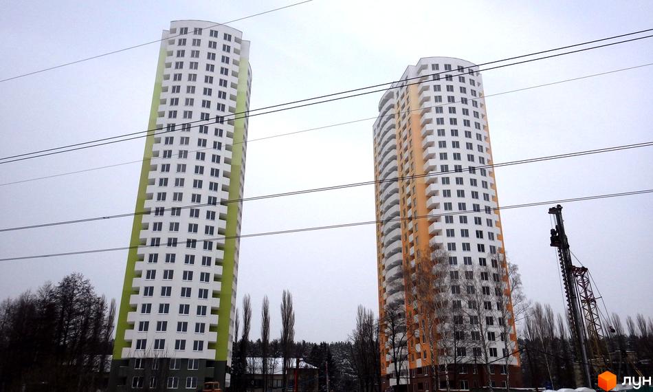 Дві висотні новобудови ЖК Озерний (Обухівська, 135а) з білими фасадами та яскравими зеленими та помаранчевими вставками. Навколо - дерева та будівельна техніка.