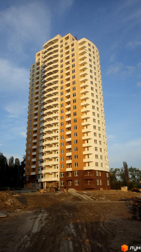 Зовнішній вигляд житлового комплексу Озерний (Обухівська, 135а) з численними балконами та вікнами на тлі блакитного неба. На передньому плані - будівельний майданчик з землею та будівельними матеріалами.