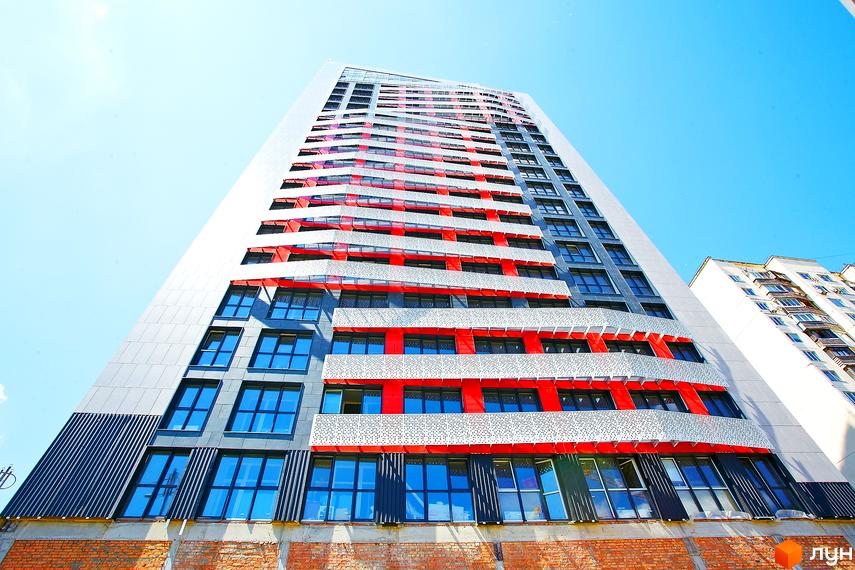Фасад житлового комплексу Квартет з червоними та білими елементами, великими вікнами та сірими декоративними панелями. Будинок має незвичайну архітектуру та виглядає вражаюче на тлі блакитного неба.