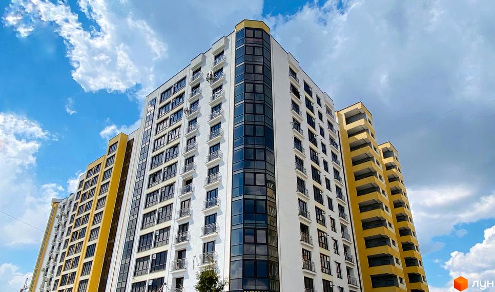 Новобудова ЖК Lystopad з біло-жовтими фасадами та панорамним склінням на фоні блакитного неба з хмарами. Зовнішній вигляд будинку з численними балконами та вікнами.