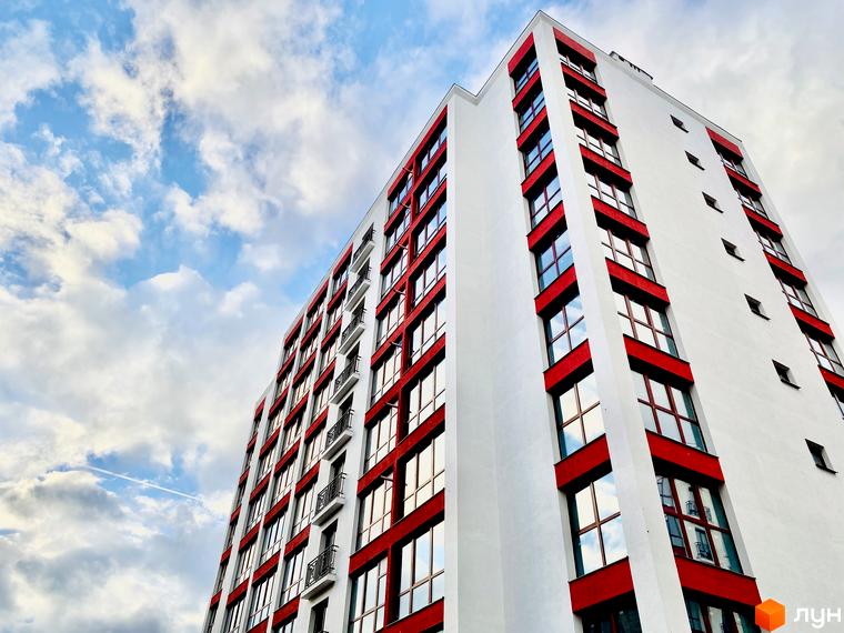Фасад Житлового комплексу Містечко Козацьке: біла багатоповерхова будівля з червоними віконними рамами на тлі блакитного неба з хмарами.