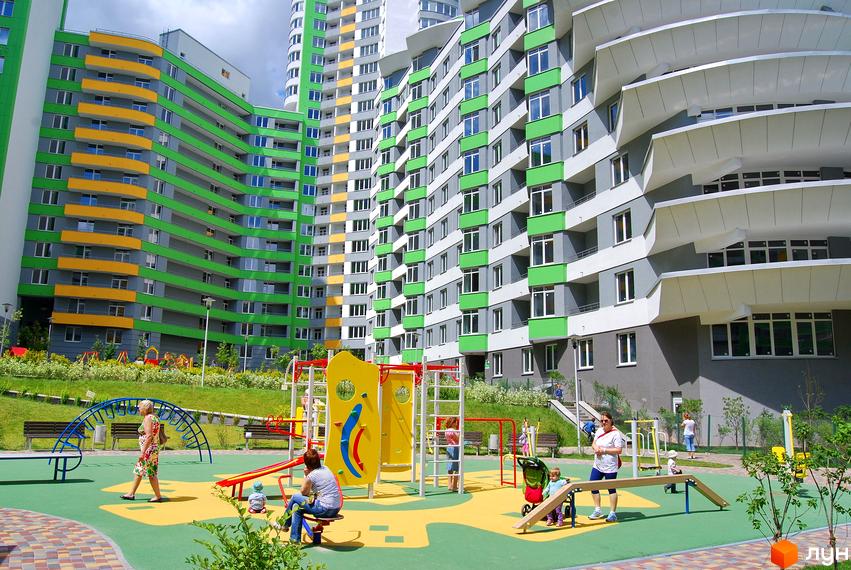 Багатоповерхові будинки Житлового комплексу Паркове місто з сірими, зеленими та жовтими фасадами. На передньому плані дитячий майданчик з яскравим обладнанням та людьми.