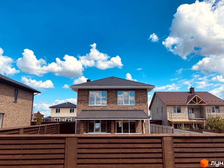 Котеджне містечко КМ Весняний: двоповерхові цегляні будинки з темними дахами на тлі блакитного неба з хмарами. Прибудинкова територія огороджена дерев'яним парканом.