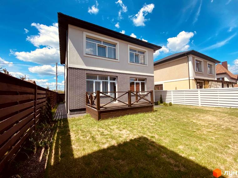 Котеджне містечко КМ Весняний. Двоповерховий будинок з цегляним цоколем та дерев'яною терасою на тлі зеленого газону та паркану.