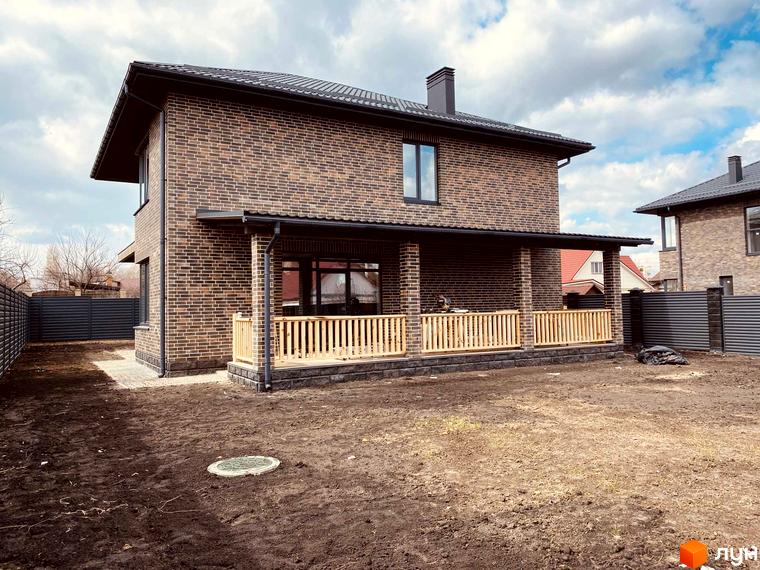 Котеджне містечко КМ Весняний: двоповерховий цегляний будинок з терасою та дерев'яною огорожею на тлі блакитного неба з хмарами. Навколо будинку облаштована прибудинкова територія з газоном та парканом.
