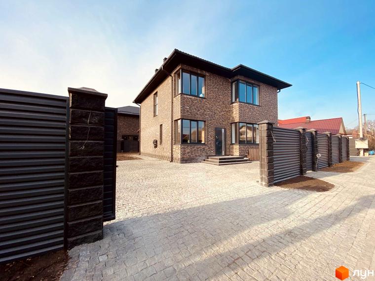 Котеджне містечко КМ Весняний: двоповерховий цегляний будинок з великими вікнами та вимощеною територією перед ним. Огороджена територія з парканом та воротами.