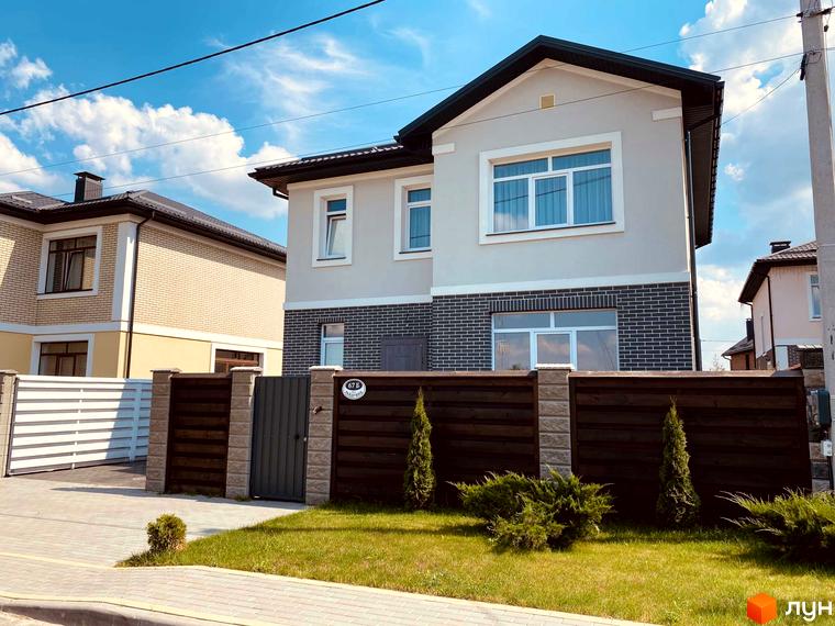Котеджне містечко Весняний: двоповерховий будинок з цегляним цоколем та дерев'яним парканом. Прибудинкова територія з газоном та зеленими насадженнями.