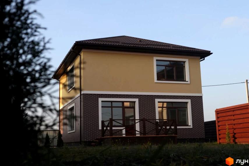 Котеджне містечко Весняний: двоповерховий будинок з бежевим фасадом, цегляним цоколем та верандою. Навколо зелень.