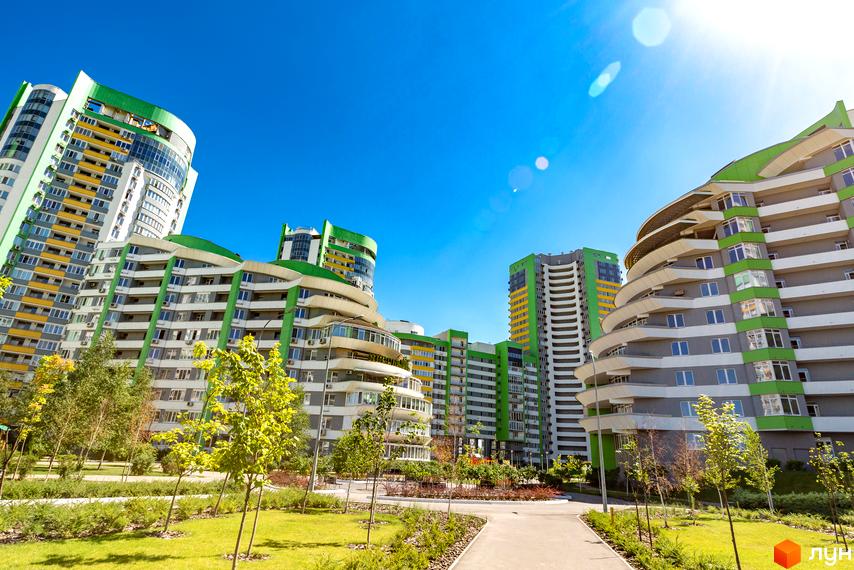 Житловий комплекс Паркове місто: багатоповерхові будинки з зеленими та сірими фасадами, вигнутими балконами. На передньому плані доглянутий газон, молоді дерева та доріжки.