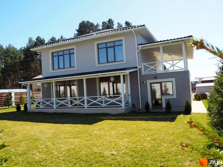 Котеджне містечко КМ Лісова Білогородка: двоповерховий сірий будинок з білою верандою та зеленим газоном. Навколо будинку розташовані дерева та доглянута прибудинкова територія.