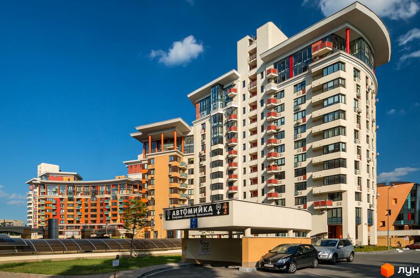 Житловий комплекс Сонячна брама з вигнутими фасадами та червоними балконами. На передньому плані видно в'їзд до підземного паркінгу та зелену зону.