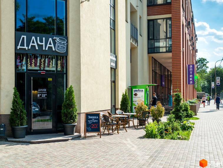 Житловий комплекс Сонячна брама: багатоповерховий будинок зі світлим фасадом та цегляними вставками. Навколо – бруківка, зелені насадження та літні столики.