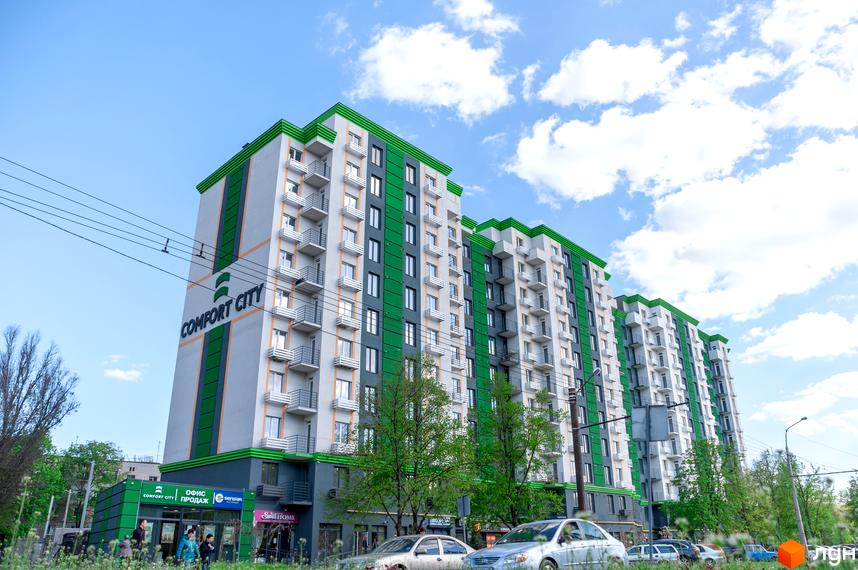 Багатоповерхові будинки Житлового комплексу Comfort City з сірими фасадами, зеленими вертикальними та горизонтальними акцентами. Навколо видно зелені дерева та автомобілі.