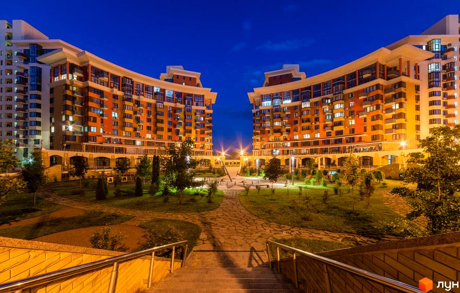 Нічний вигляд житлового комплексу Сонячна брама з двома вигнутими будинками з помаранчевими фасадами та освітленим внутрішнім двором з зеленими насадженнями.