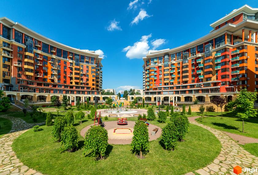 Два вигнуті будинки Житлового комплексу Сонячна брама з помаранчевими фасадами та балконами. На передньому плані зелений двір з доріжками та дитячим майданчиком.