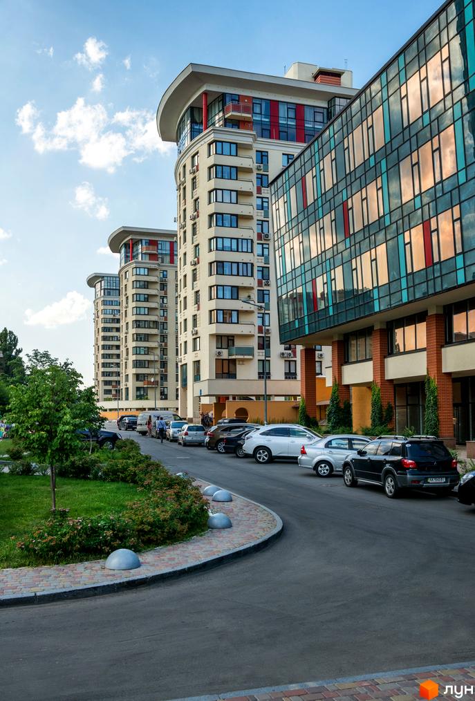 Житловий комплекс Сонячна брама: високі будинки світлого кольору з вигнутими дахами та скляним фасадом. Поруч дорога, парковка та зелені насадження.