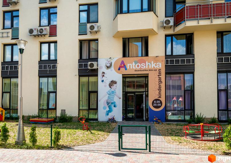 Світлий фасад будинку Житлового комплексу Сонячна брама з великими вікнами. На першому поверсі розташований дитячий садок Antoshka з ігровим майданчиком.