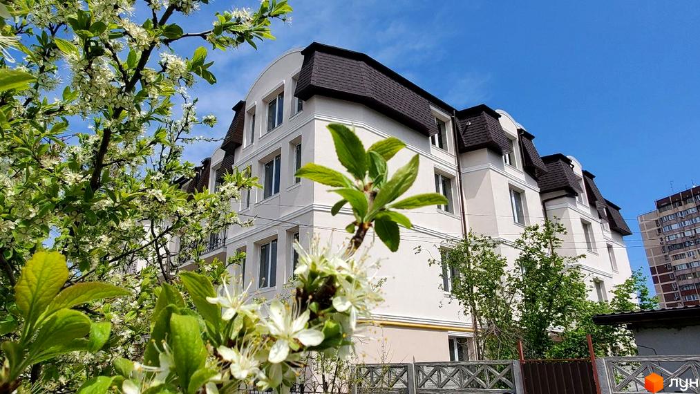 Зображення фасаду будинку Житловий комплекс вул. Академіка Вернадського (Тульська), 21а, з білими стінами та темним дахом, на фоні блакитного неба. Поруч квітучі дерева.