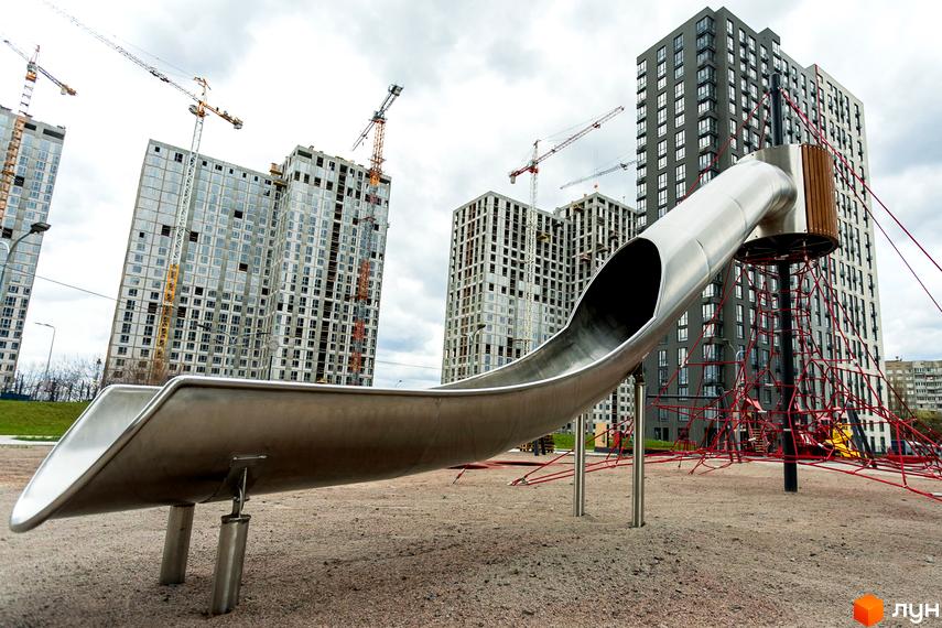 Будівельний проєкт ЖК Варшавський Плюс: висотні будинки на стадії будівництва, дитячий майданчик з металевою гіркою та червоними сітками.