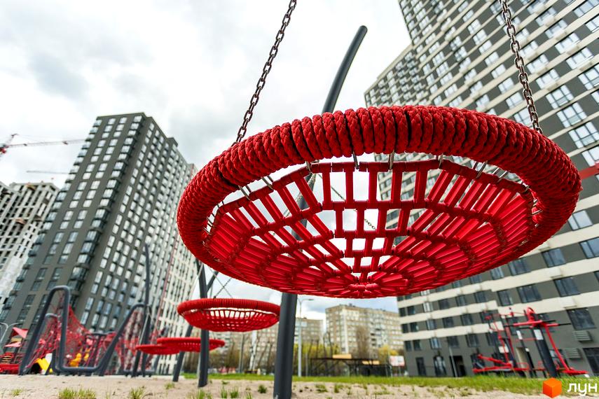 Новобудова ЖК Варшавський Плюс з червоними дитячими гойдалками на прибудинковій території. Багатоповерхові будинки на фоні хмарного неба.