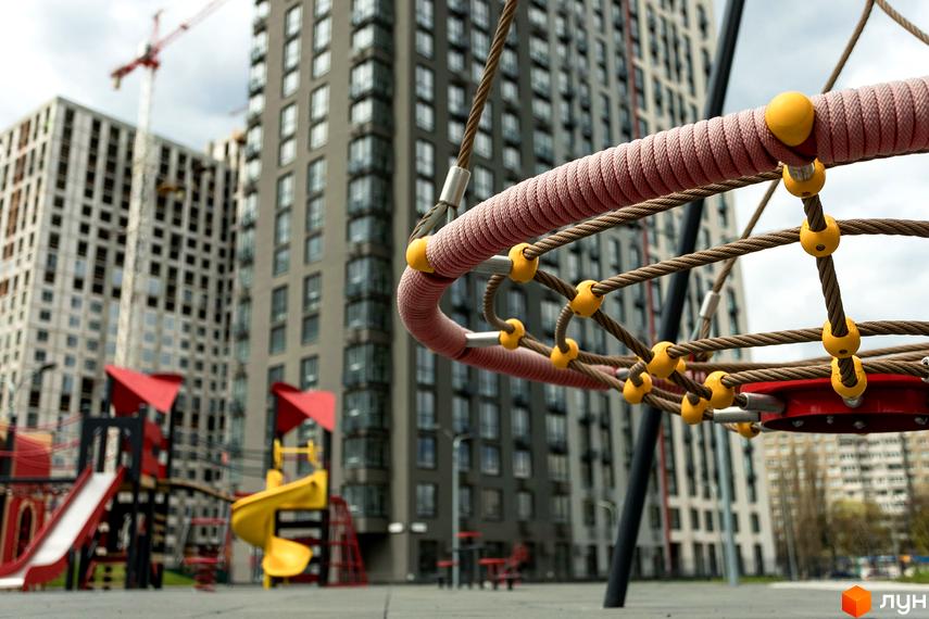 Дитячий майданчик біля багатоповерхових будинків житлового комплексу Варшавський Плюс. На передньому плані мотузковий ігровий елемент з жовтими деталями.