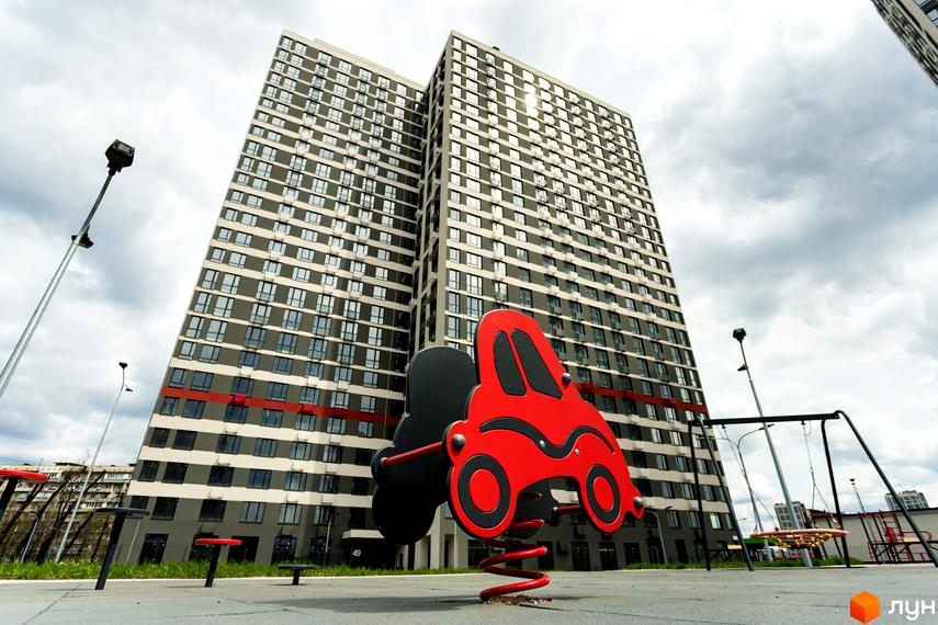 Новобудова ЖК Варшавський Плюс з дитячим майданчиком. Багатоповерхові будинки з сірими фасадами та червоними смугами.