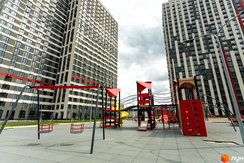 Житловий комплекс Варшавський Плюс: високі будинки з сірими та бежевими фасадами, дитячий майданчик з гірками та гойдалками.