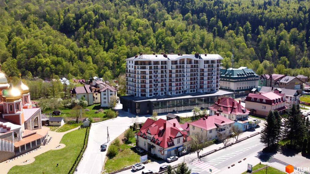 Житловий комплекс Скеля: багатоповерховий будинок білого кольору з балконами та великими вікнами на першому поверсі, розташований серед зелених гір, поруч з церквою.