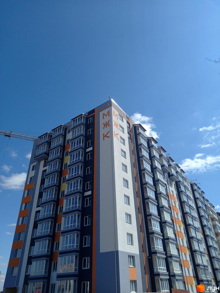 Багатоповерховий будинок МЖК Сім'я з синьо-оранжево-білим фасадом та заскленими балконами під блакитним небом. Поруч видно будівельний кран.