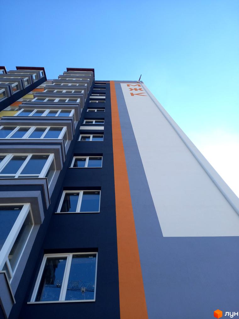 Фасад новобудови МЖК Сім'я з вікнами та балконами, виконаний у темно-синіх, помаранчевих та світло-сірих тонах на тлі блакитного неба.