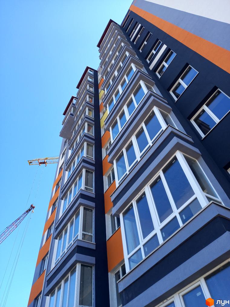 Багатоповерховий будинок Комплекс Сім'я з темно-синім, сірим та помаранчевим фасадом, великими вікнами. На тлі блакитного неба видно будівельні крани.